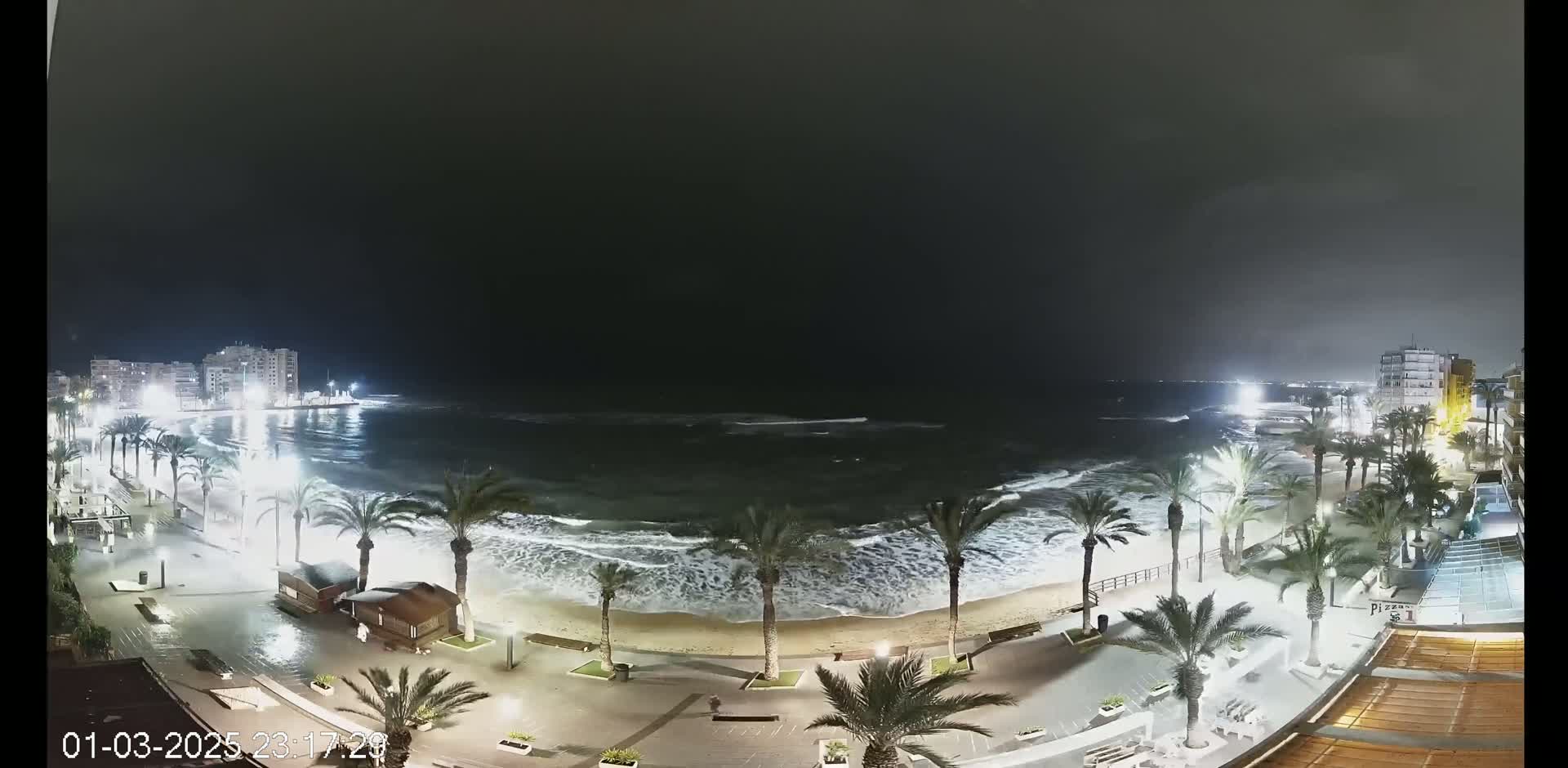 A nighttime beach scene shows moderately rough waves lapping a sandy shore, lined with palm trees and illuminated buildings under a dark, overcast sky.