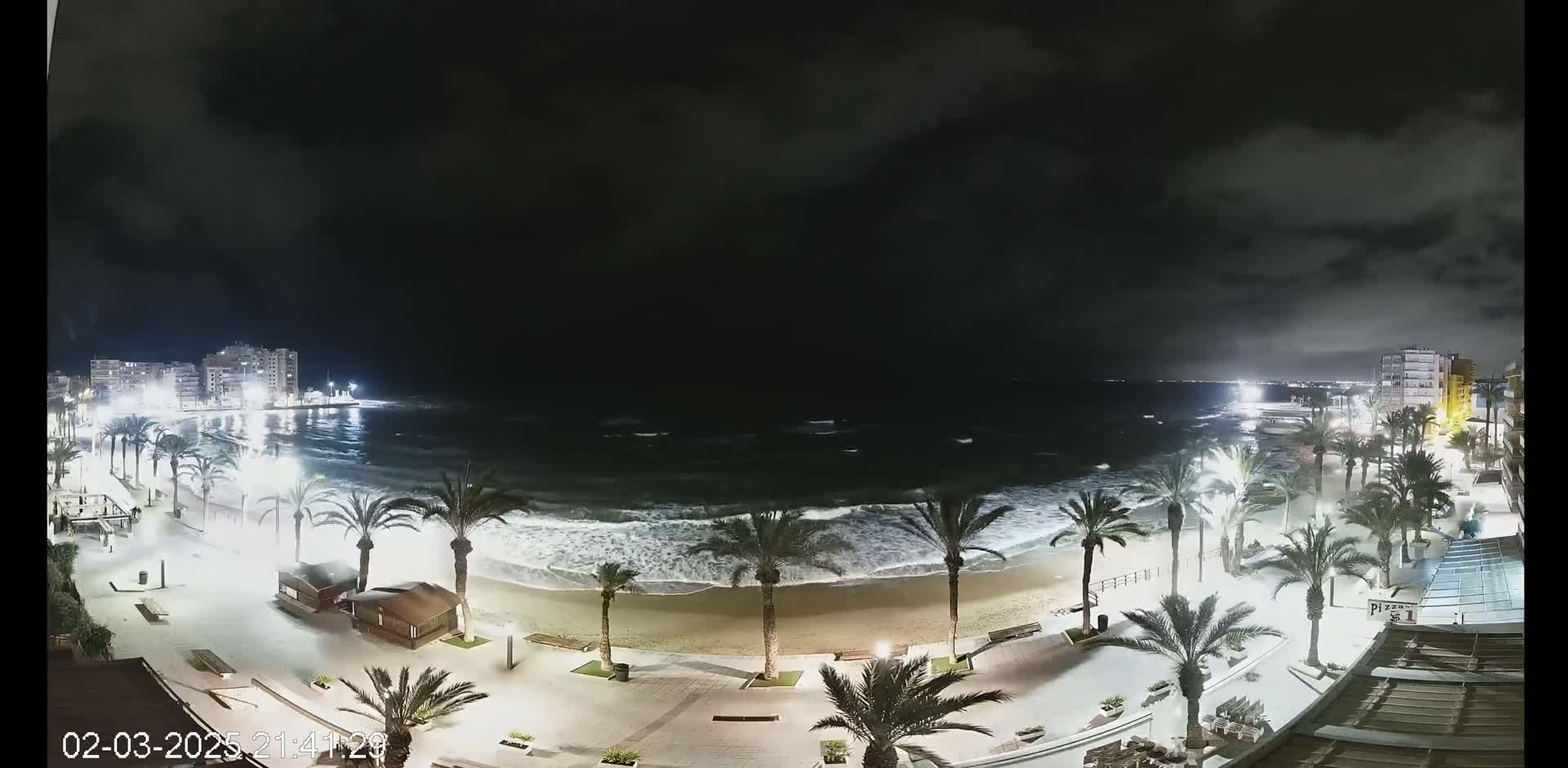 A nighttime view of a beach with palm trees lining a paved walkway, showing moderately rough waves under a cloudy sky.