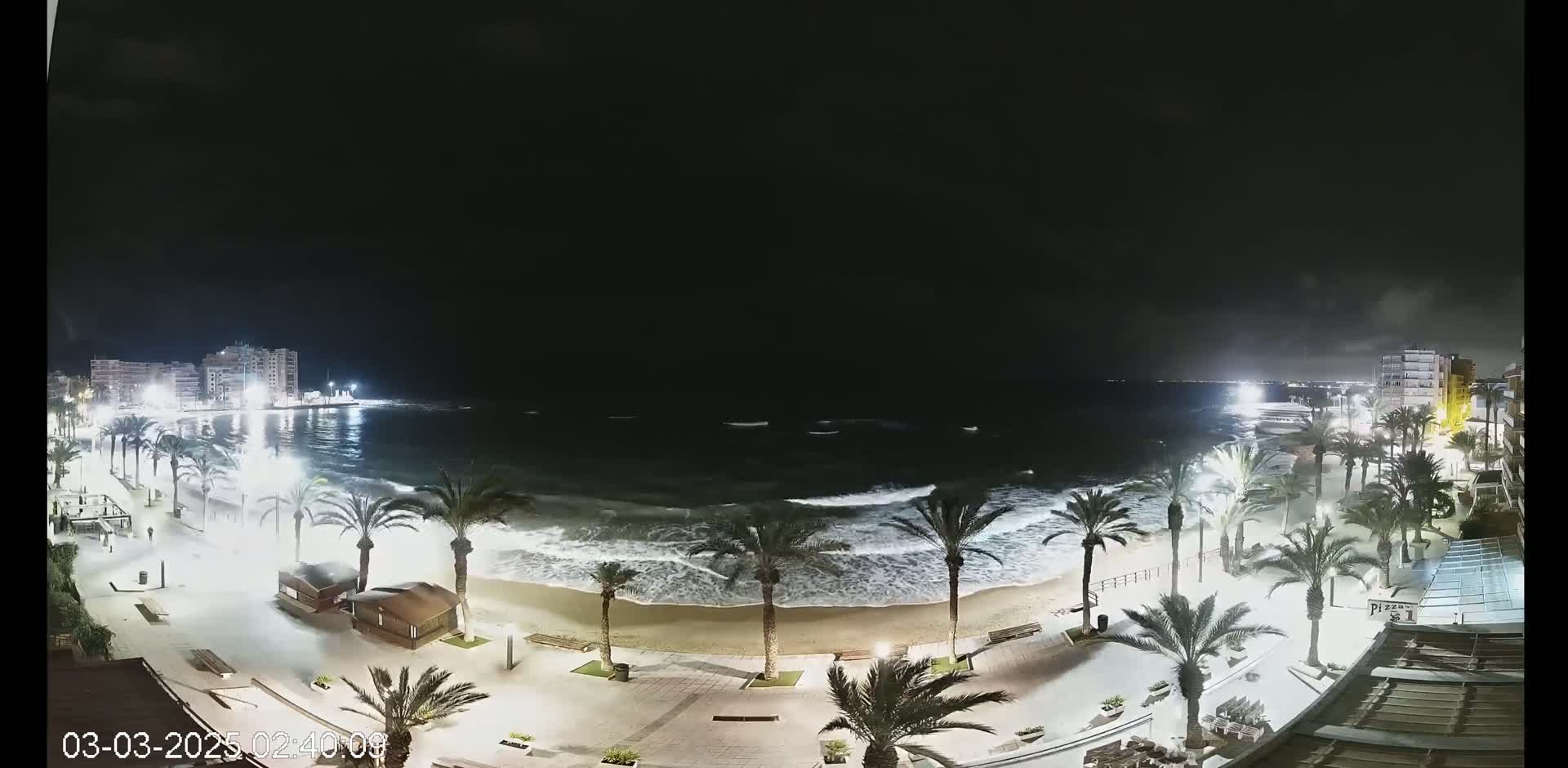 A nighttime view of a beach with moderately sized waves crashing on the shore, palm trees lining the boardwalk, and buildings visible in the distance under a dark, mostly clear sky.