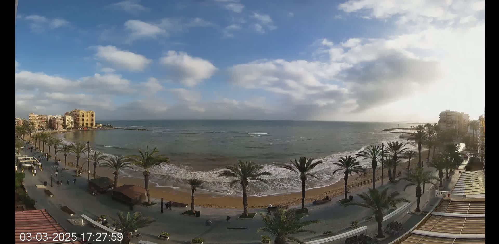 A partly cloudy day shows a beach with palm trees lining a paved walkway, buildings in the background, and gently rolling waves.