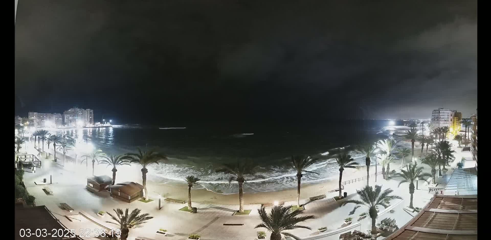 A nighttime beach scene shows moderately sized waves lapping a sandy shore lined with palm trees and illuminated walkways, under a dark, cloudy sky.