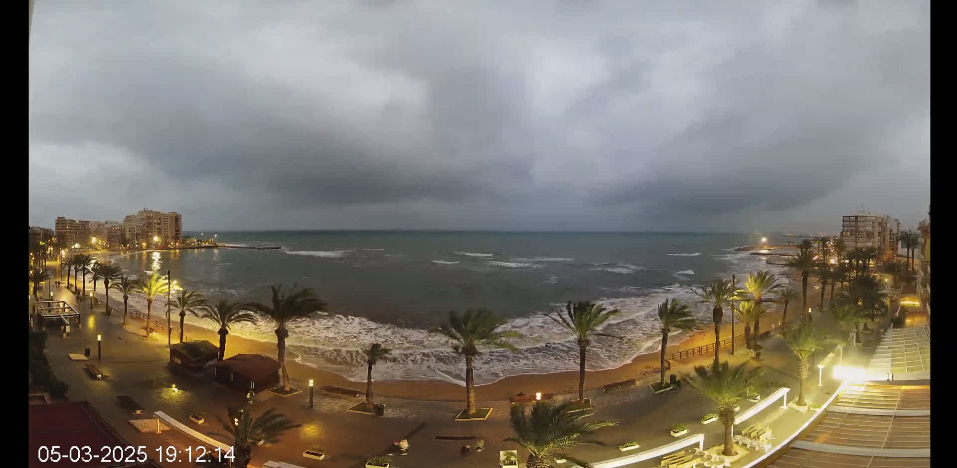 Under a dark, cloudy sky, moderately sized waves crash on a sandy beach lined with palm trees and illuminated buildings.