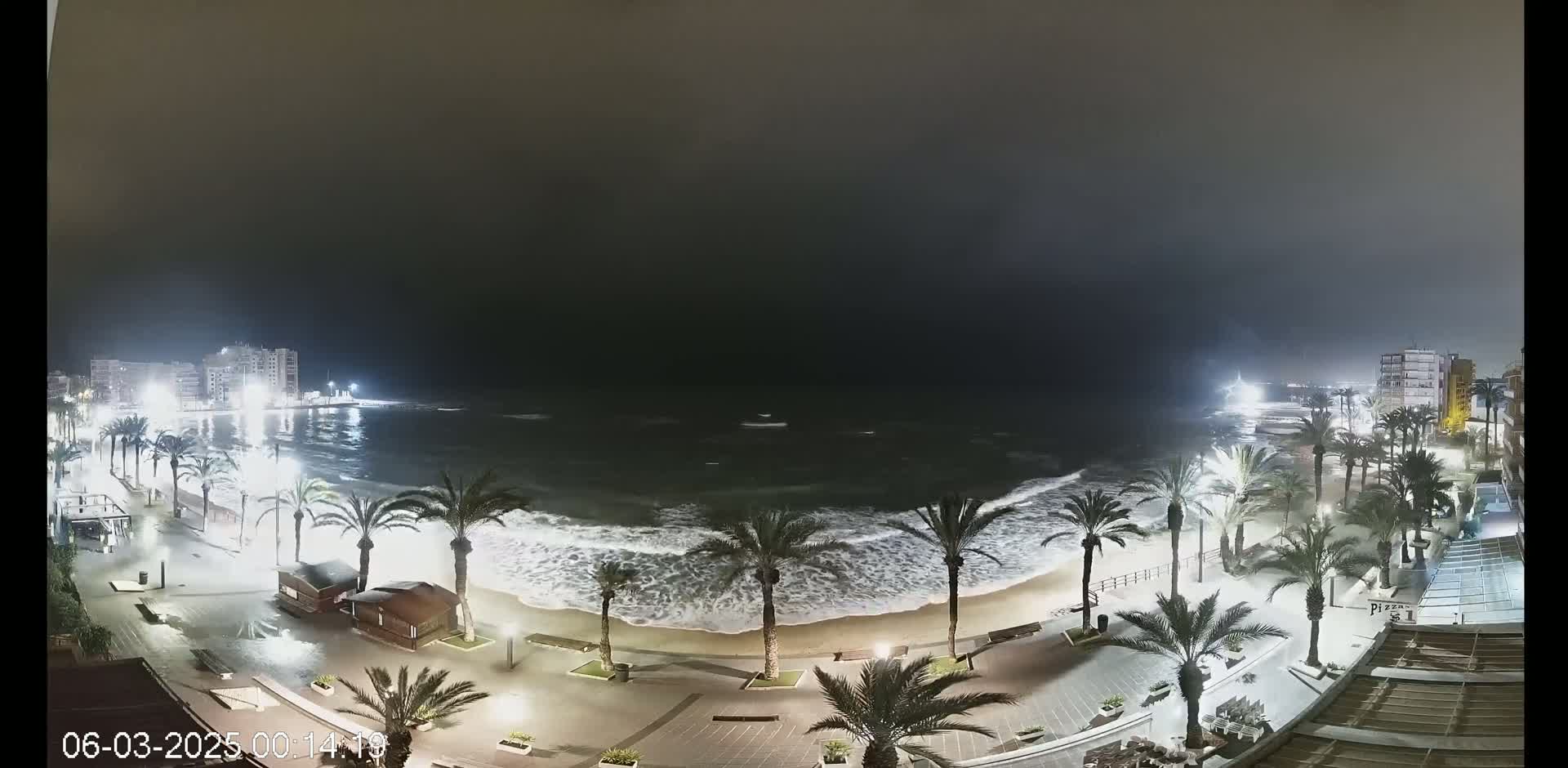 A nighttime coastal scene shows palm trees lining a paved beachfront promenade with buildings in the background, under dark and stormy conditions with large waves crashing on the shore.