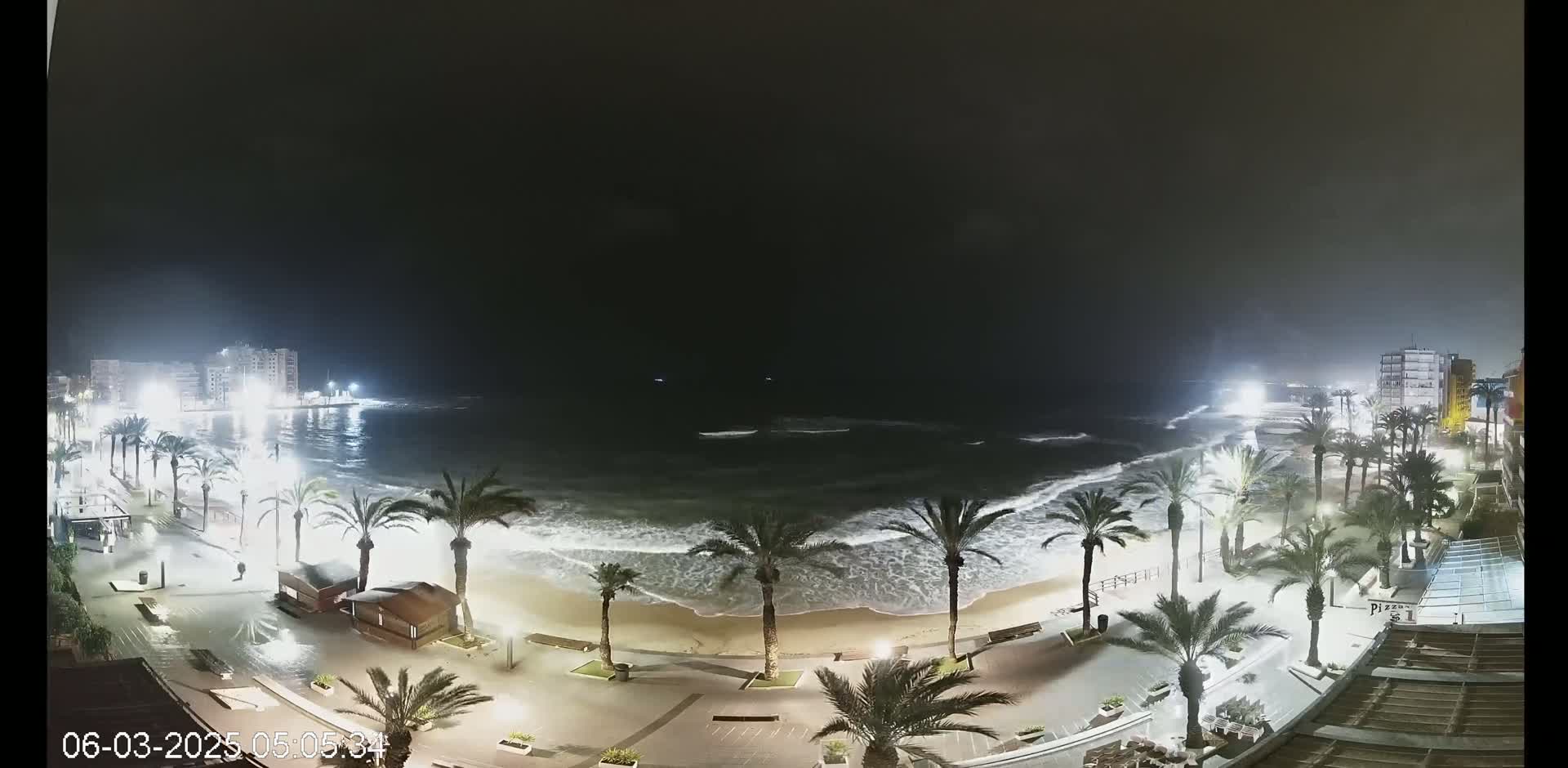 A nighttime view of a beach at high tide, with palm trees lining a paved walkway and buildings visible in the background under a dark, cloudy sky.