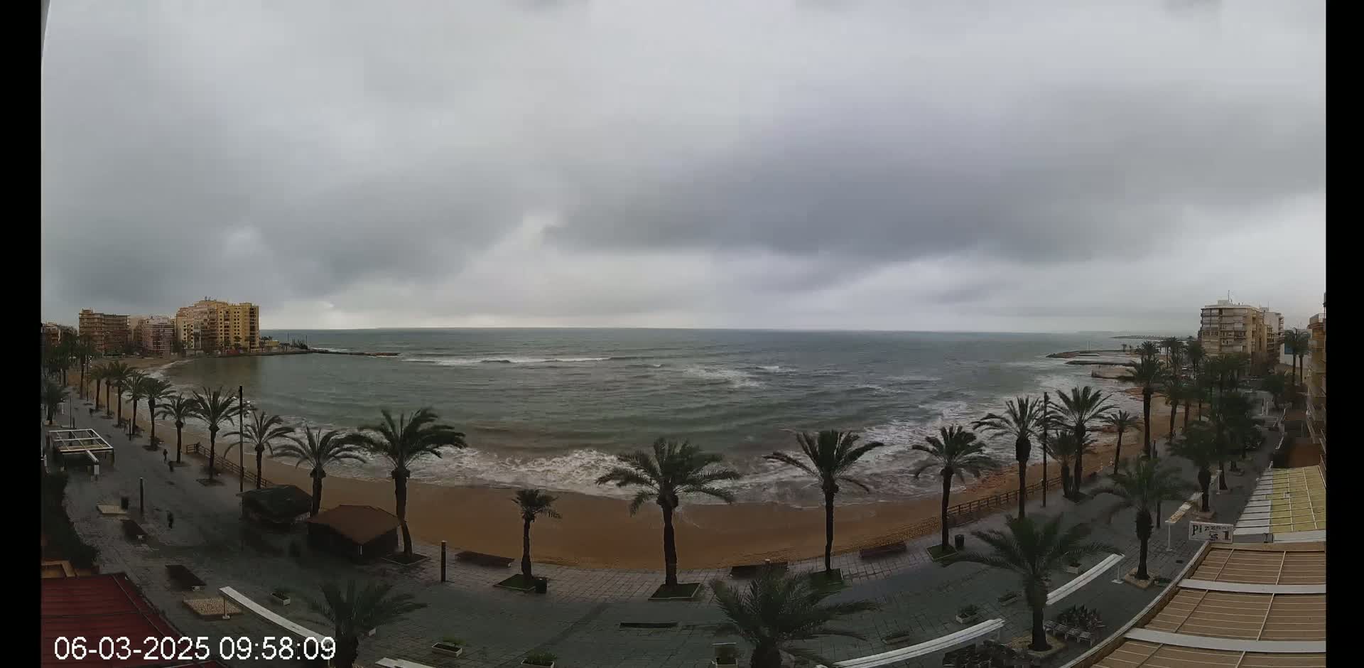 A sandy beach with palm trees lines a promenade next to a wavy, grey ocean under an overcast sky.