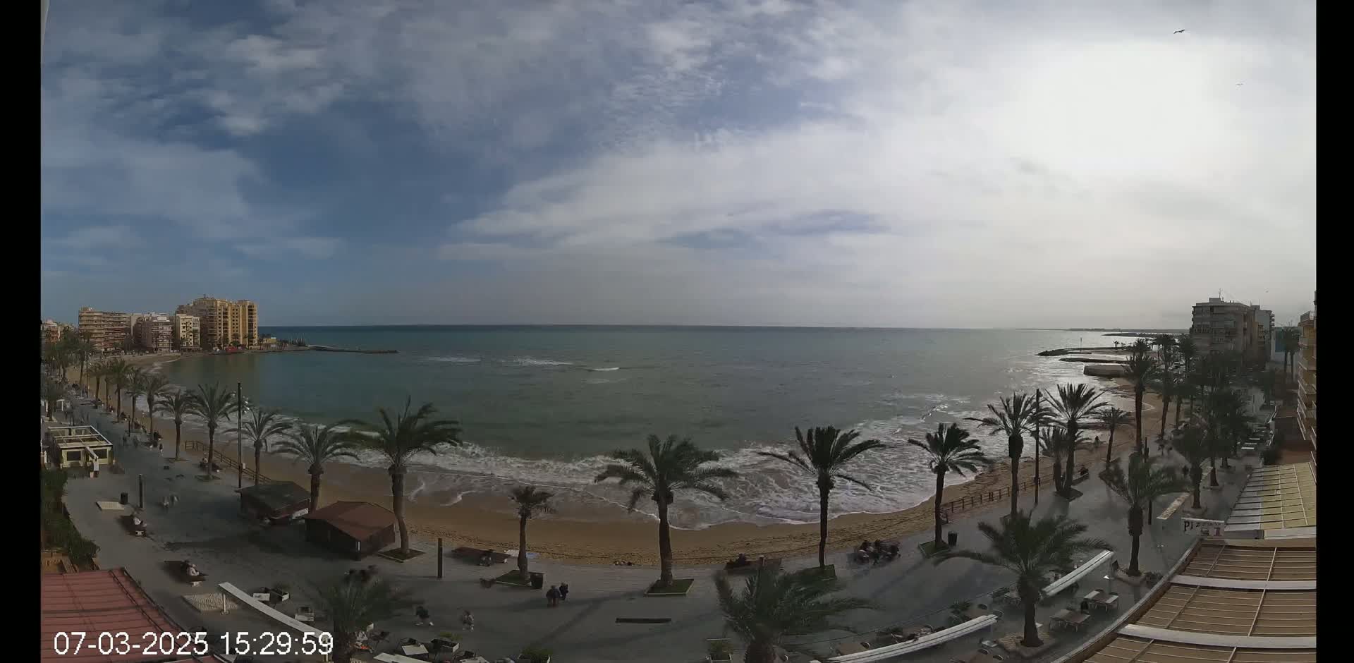 A mostly cloudy day shows a sandy beach with palm trees lining a paved walkway, overlooking a calm sea with small waves and buildings in the distance.