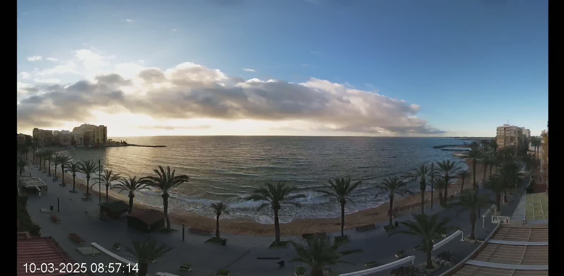 A partly cloudy sunrise illuminates a beach with palm trees lining a promenade, overlooking a calm sea with gentle waves.