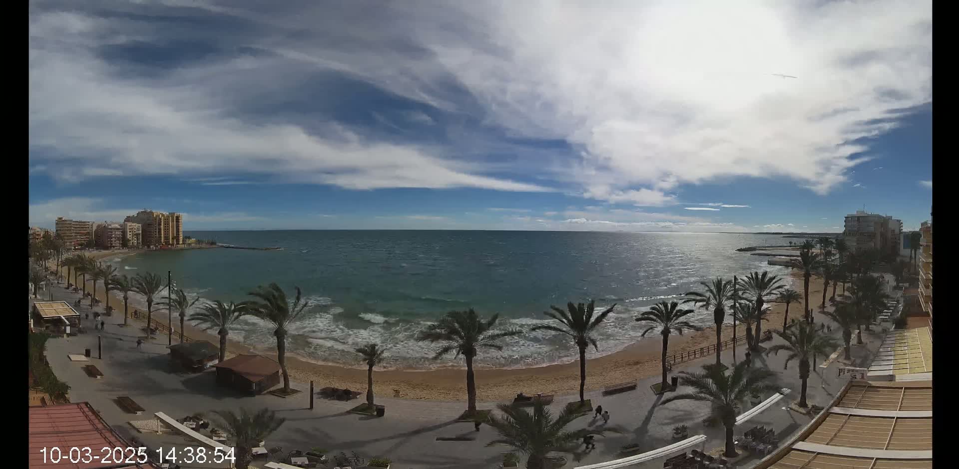 A partly cloudy day shows a sandy beach with palm trees lining a paved walkway, overlooking a moderately wavy ocean with buildings in the distance.