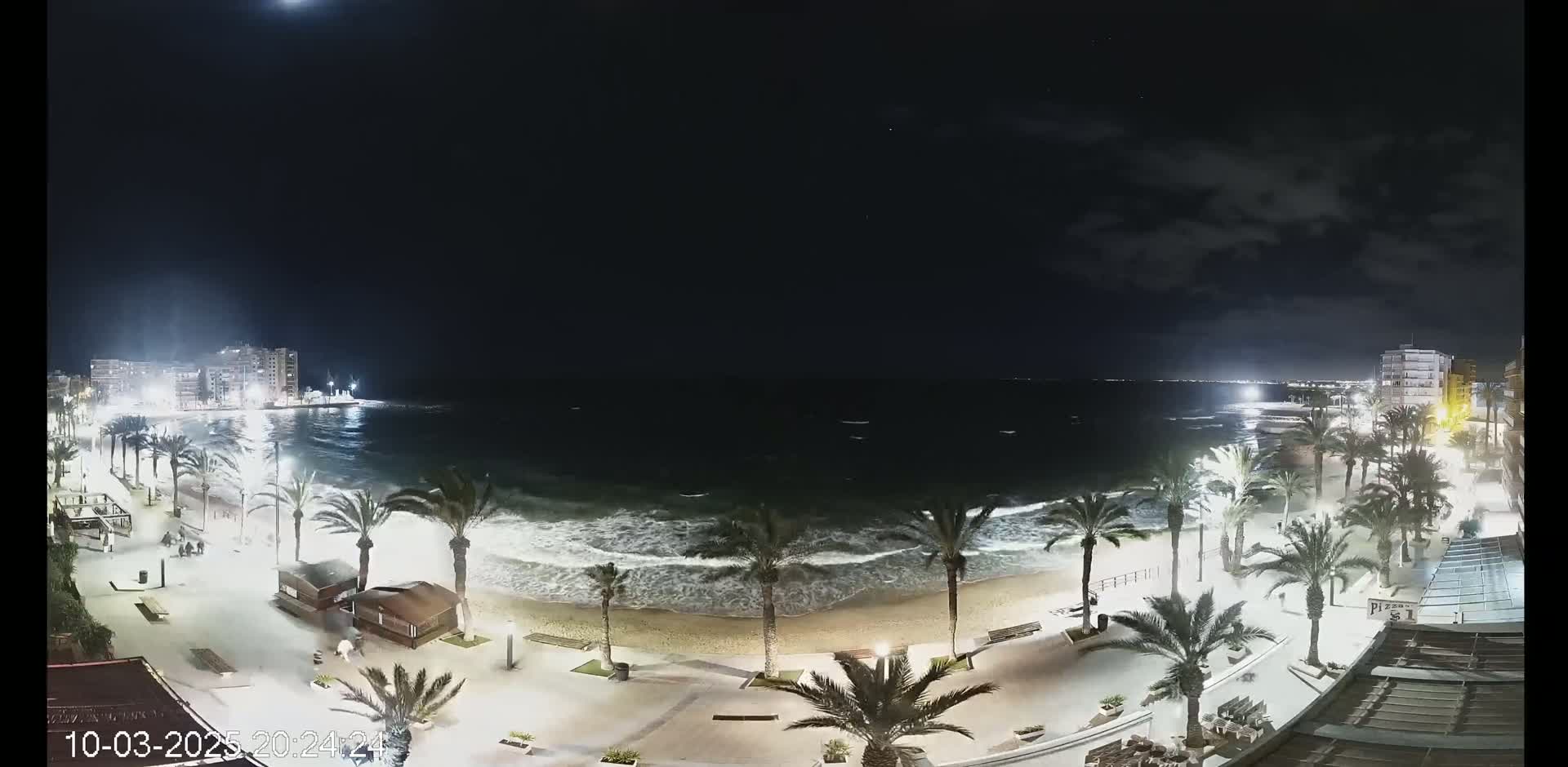 A nighttime view of a beach with palm trees, buildings in the distance, and moderately rough waves under a partly cloudy sky.