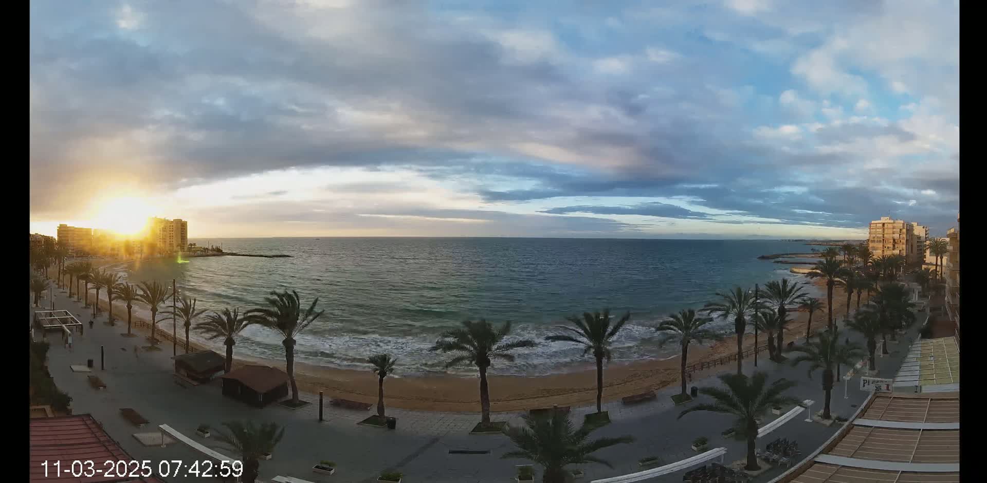 A sunrise over a calm ocean beach, palm trees lining a paved walkway next to the sand, with buildings visible in the distance under a partly cloudy sky.