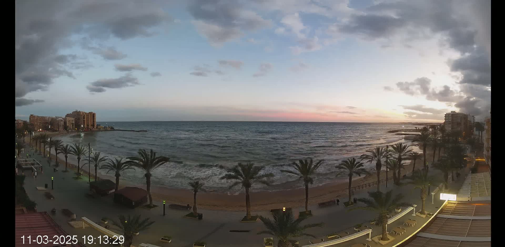 A beach with palm trees lining a paved walkway, overlooks a moderately wavy ocean under a partly cloudy twilight sky.