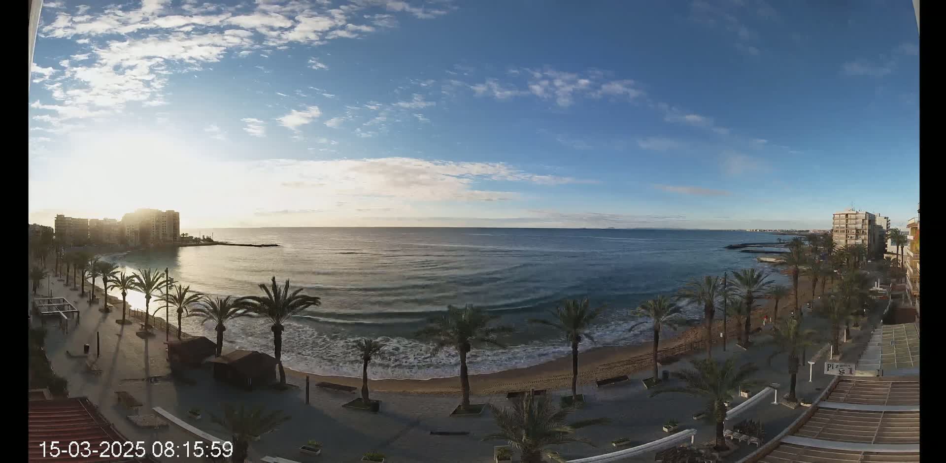 A mostly sunny day with light clouds over a sandy beach lined with palm trees and buildings, showing a calm sea with small waves.
