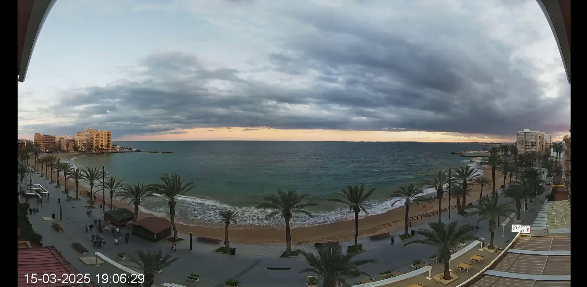 A beachfront promenade lined with palm trees overlooks a calm sea under a cloudy sunset sky.