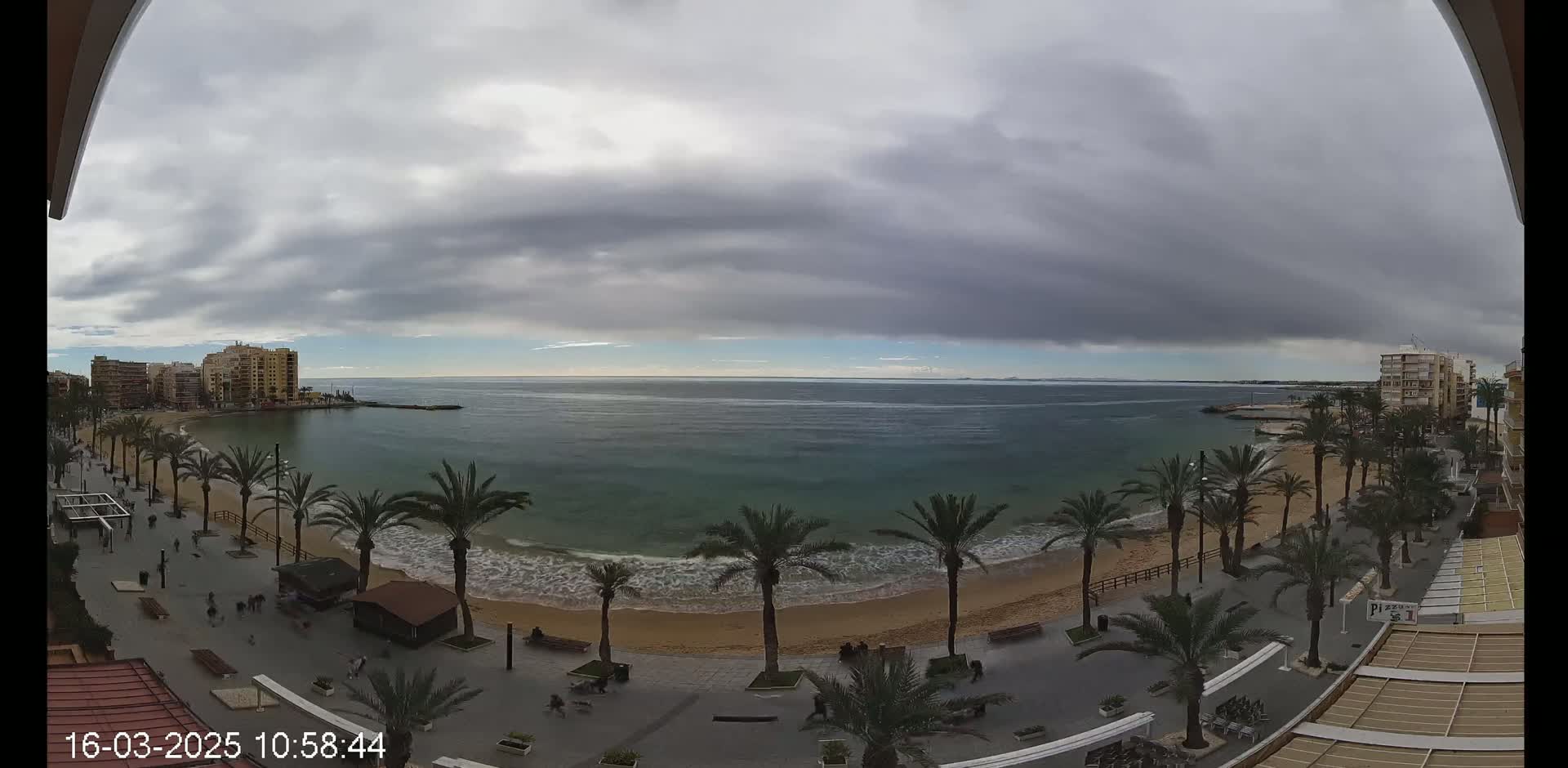 A mostly cloudy day overlooks a sandy beach lined with palm trees and buildings, with calm waves lapping the shore.