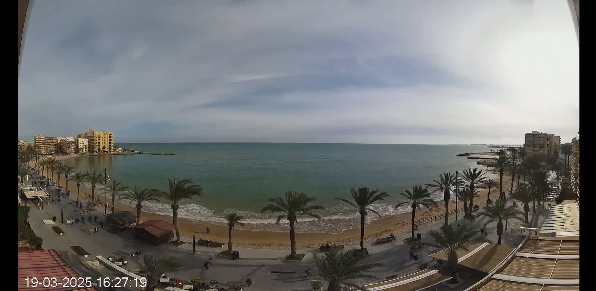 A mostly cloudy day reveals a beach with palm trees lining a paved walkway and buildings in the distance, overlooking a calm sea.