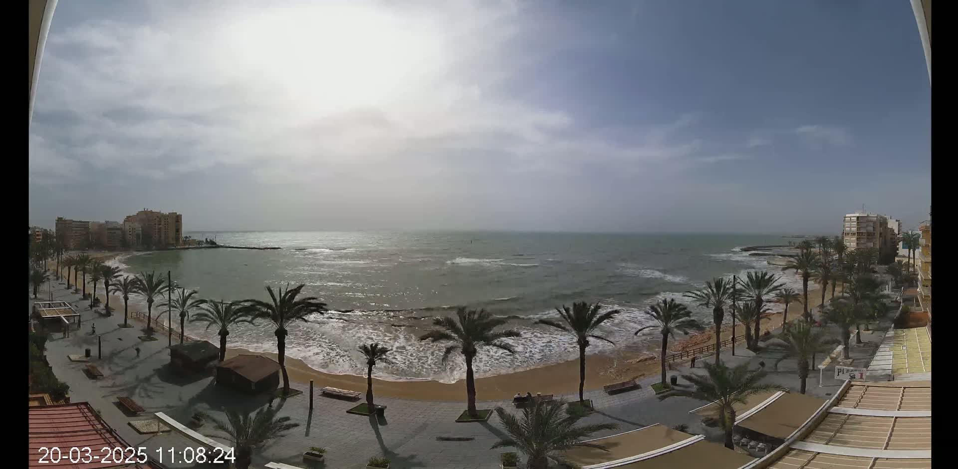 A partly cloudy day shows a beachfront view with palm trees lining a paved walkway next to a sandy beach with moderate waves.