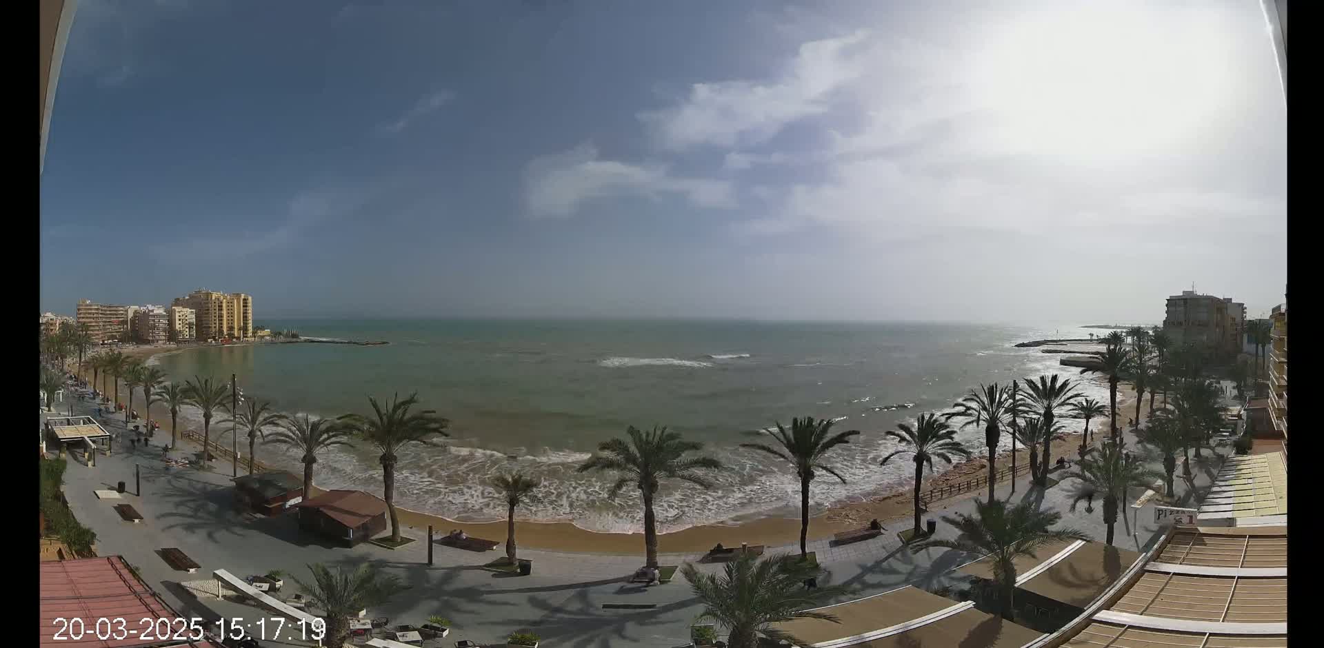 A partly cloudy day shows a beach with palm trees lining a paved walkway next to a calm, somewhat choppy ocean with small waves.