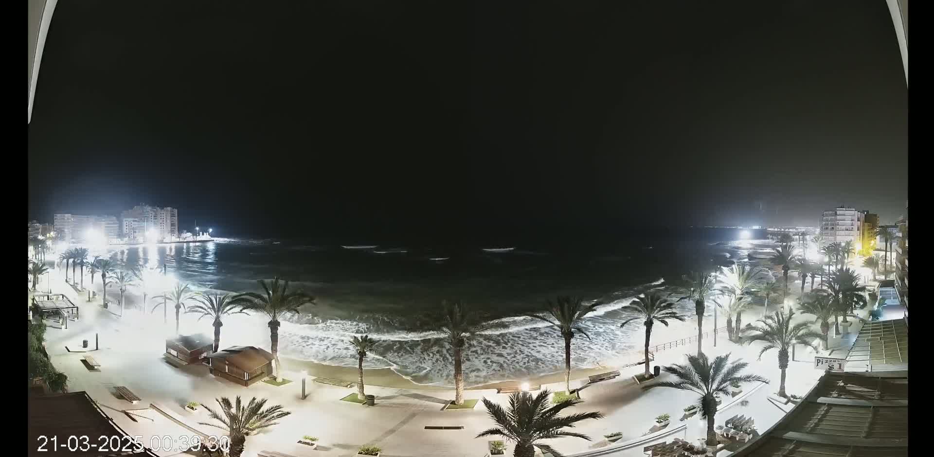 A nighttime beach scene shows moderate waves crashing on the shore under a dark sky, with palm trees lining a paved walkway next to buildings illuminated by lights.