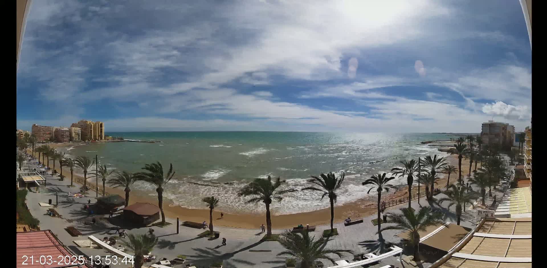 A partly sunny day shows a wavy ocean meeting a sandy beach lined with palm trees and a paved walkway; buildings are visible in the background.