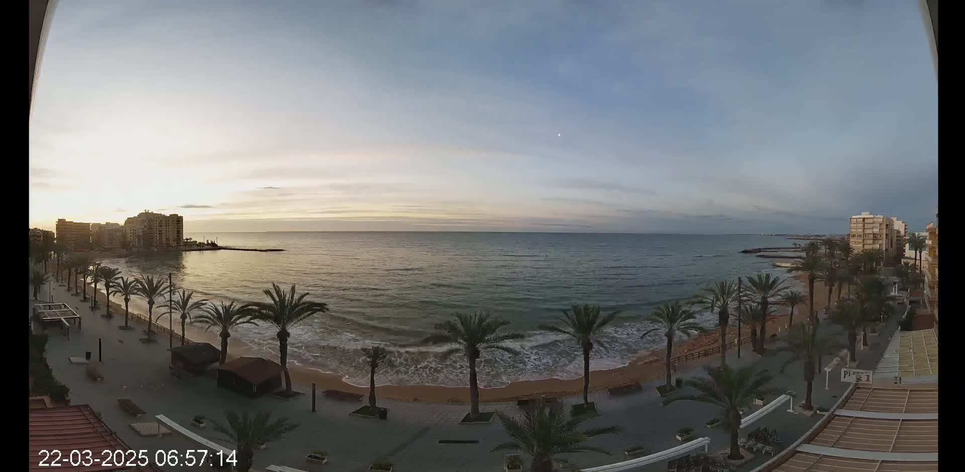 A sunrise over a calm sea with gentle waves lapping a sandy beach lined with palm trees and buildings in the background.