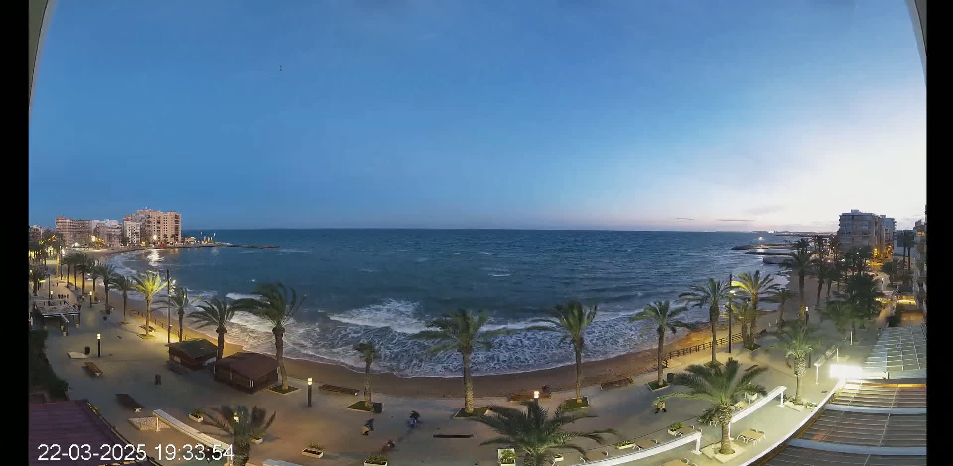 A seaside promenade at dusk with palm trees, some people walking, and moderately wavy water under a clear blue sky.