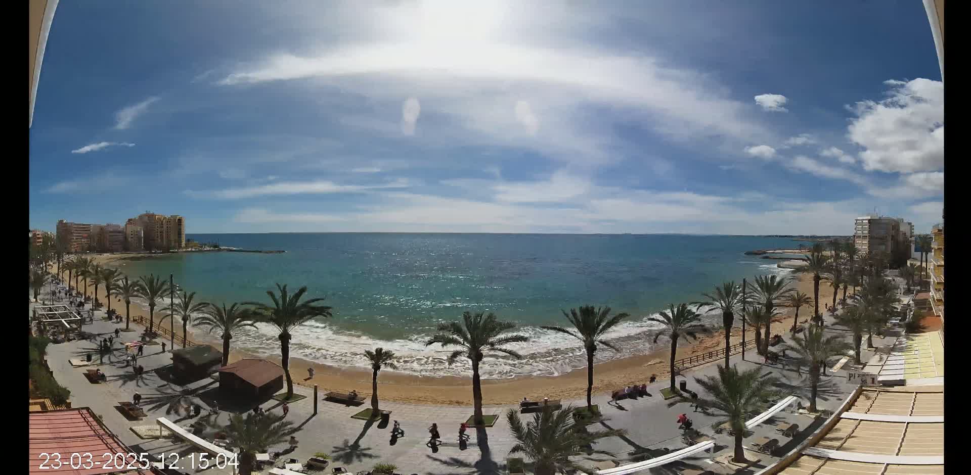 A sunny day reveals a sandy beach lined with palm trees, a boardwalk with people strolling, and a calm sea with buildings visible in the distance.