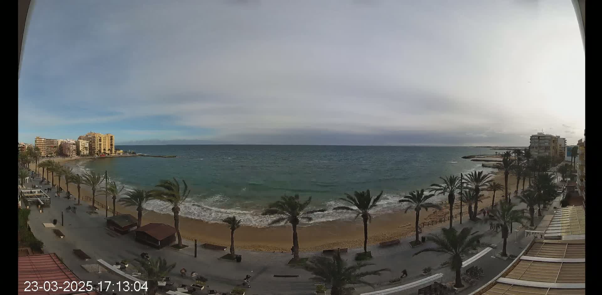 A sandy beach with palm trees lining a paved walkway, overlooking a somewhat choppy sea under a mostly cloudy sky.