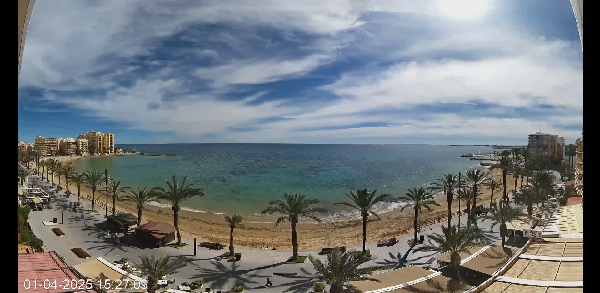 A mostly sunny day shows a sandy beach lined with palm trees and buildings, next to a calm, teal ocean.