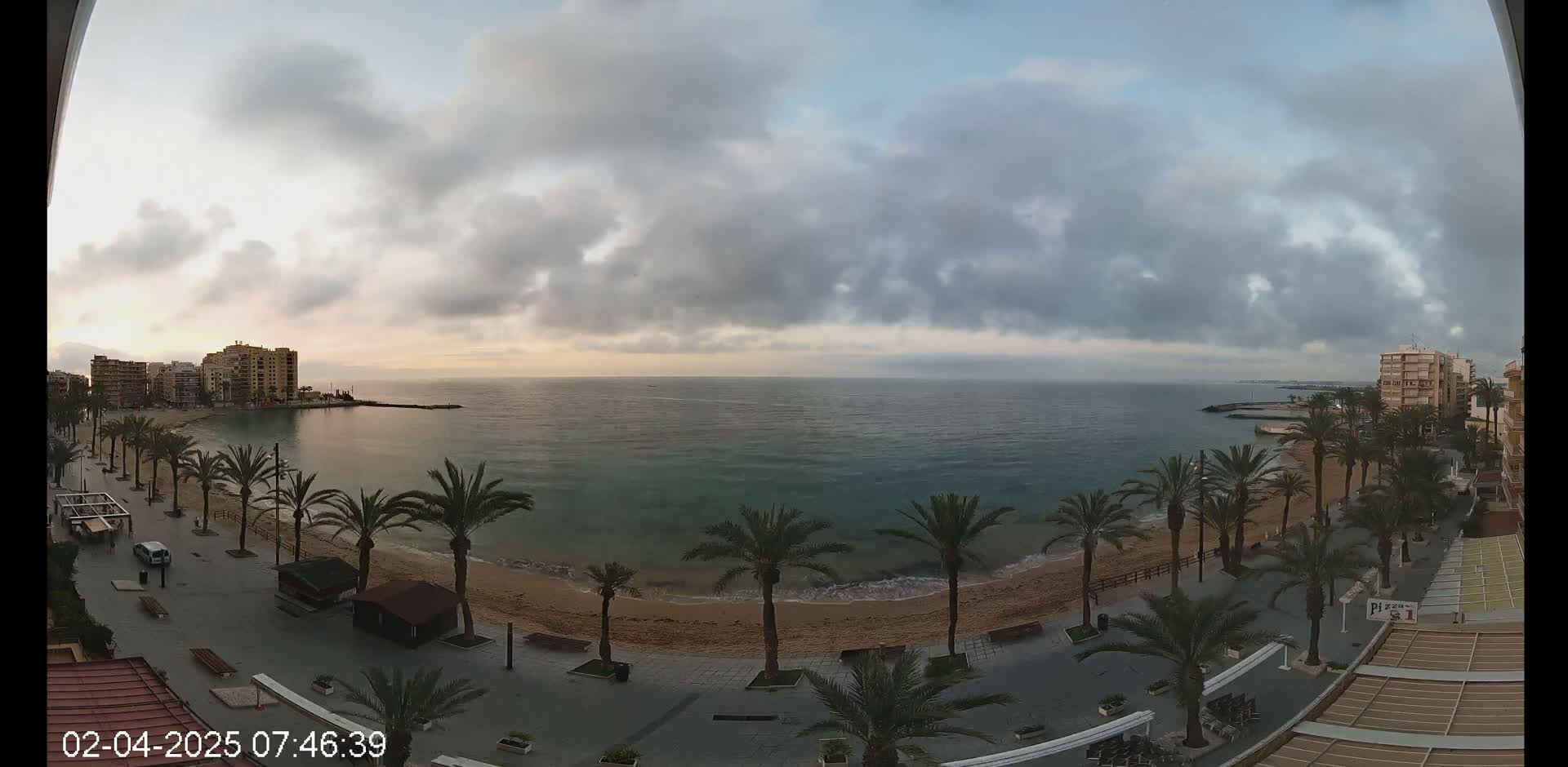 A mostly cloudy sunrise over a calm sea, with a sandy beach lined with palm trees and buildings visible in the distance.