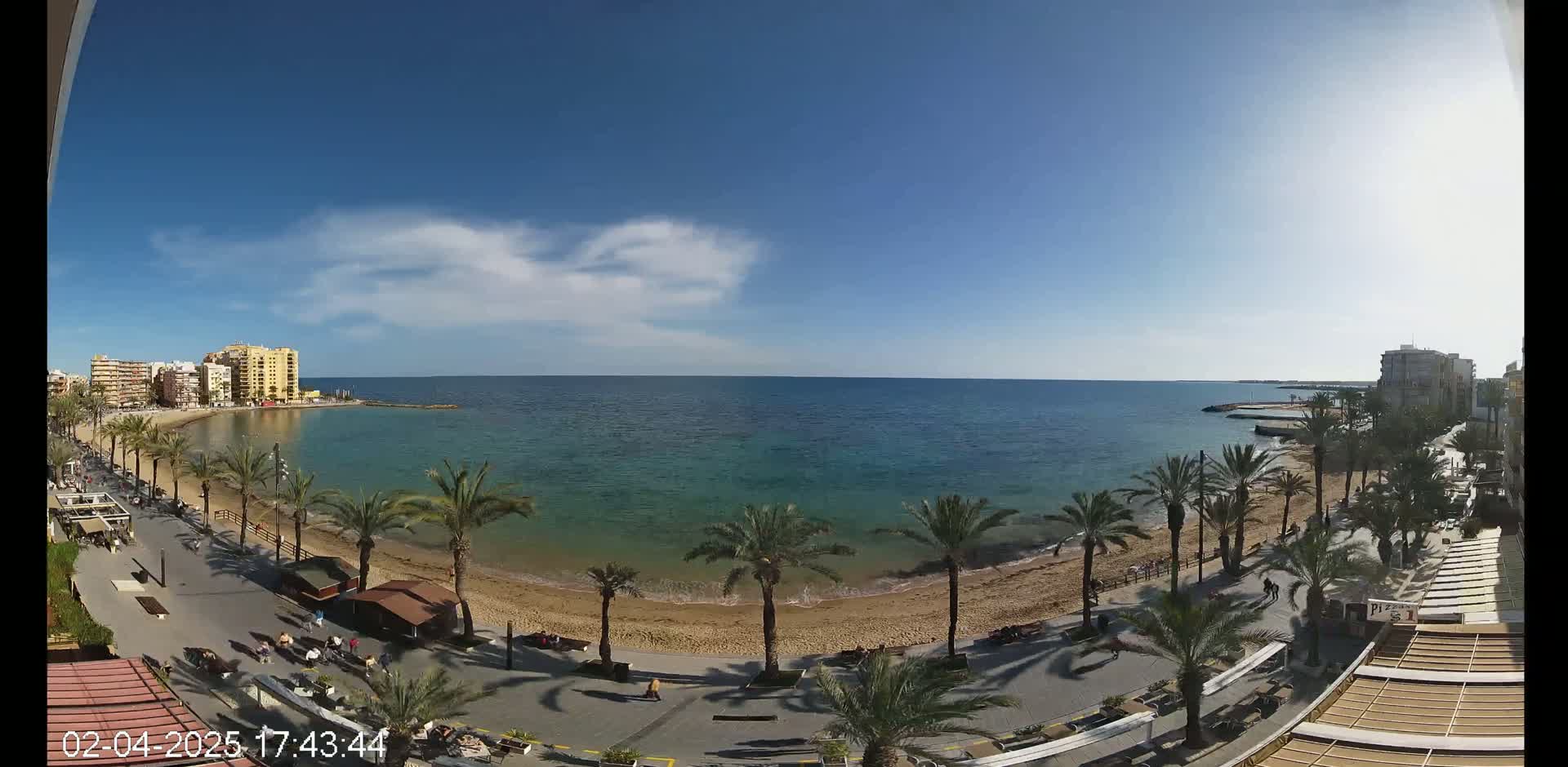 A sandy beach with palm trees lines a paved boardwalk next to a calm, teal ocean under a mostly sunny sky with a few scattered clouds.