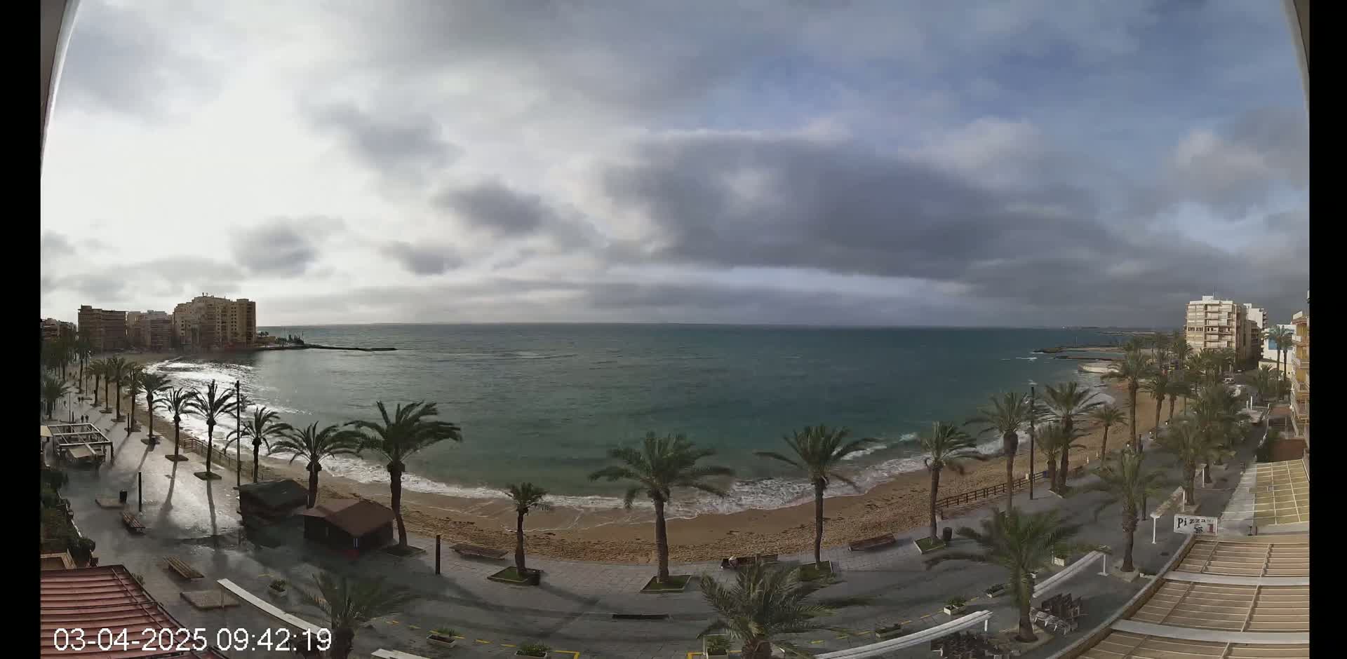 A mostly cloudy day reveals a sandy beach lined with palm trees and buildings along a calm sea.