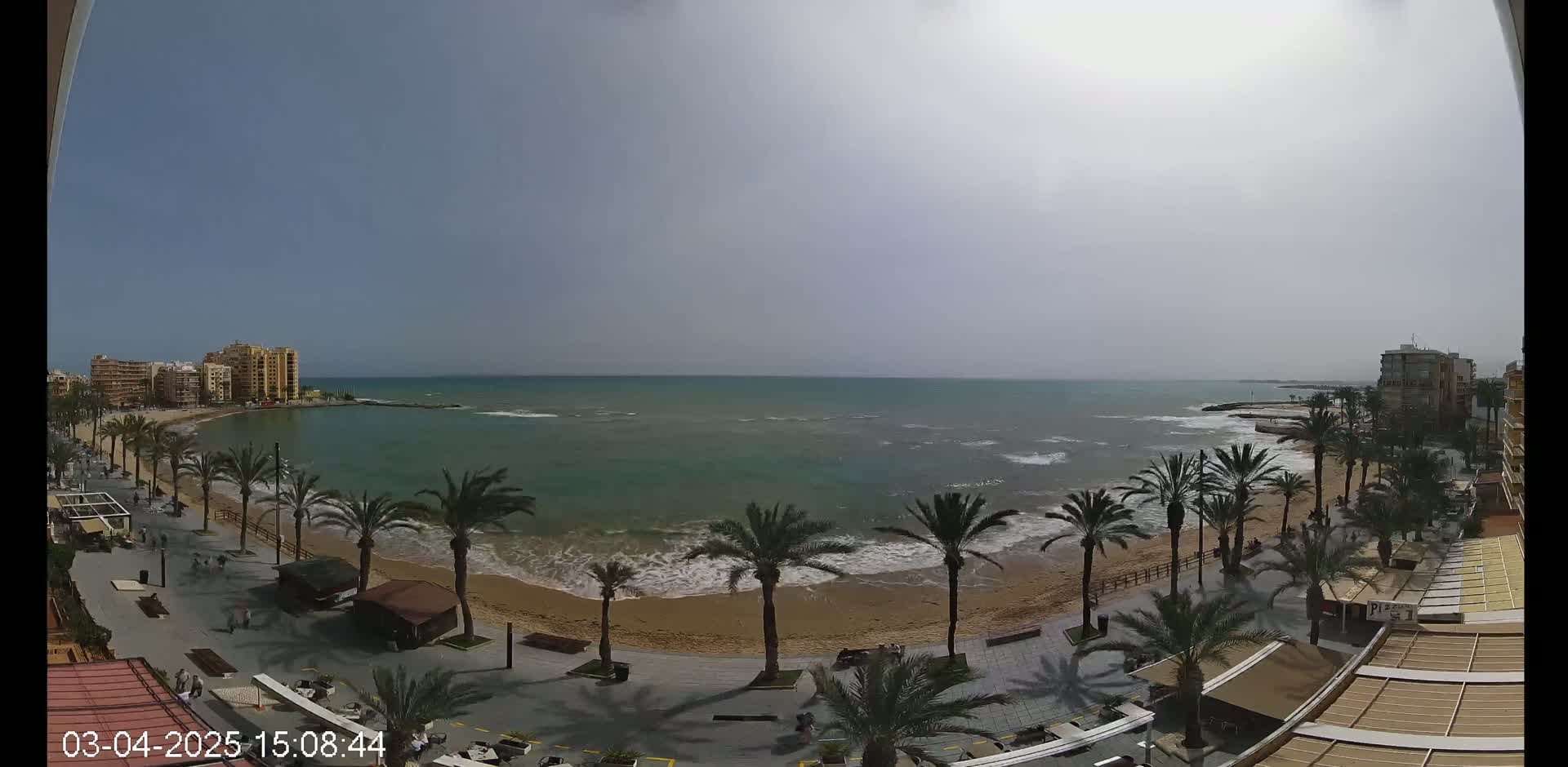 A sandy beach with palm trees lining a boardwalk, overlooks a calm sea under a hazy sky.