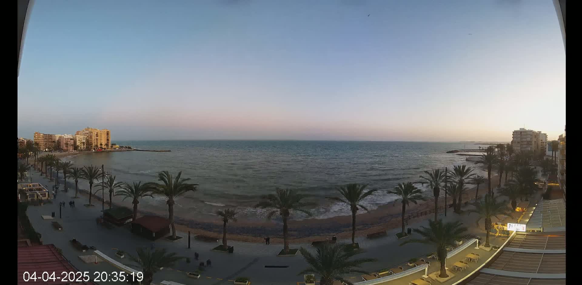 A beachfront promenade at dusk, lined with palm trees and buildings, overlooks a calm sea with small waves under a clear, twilight sky.
