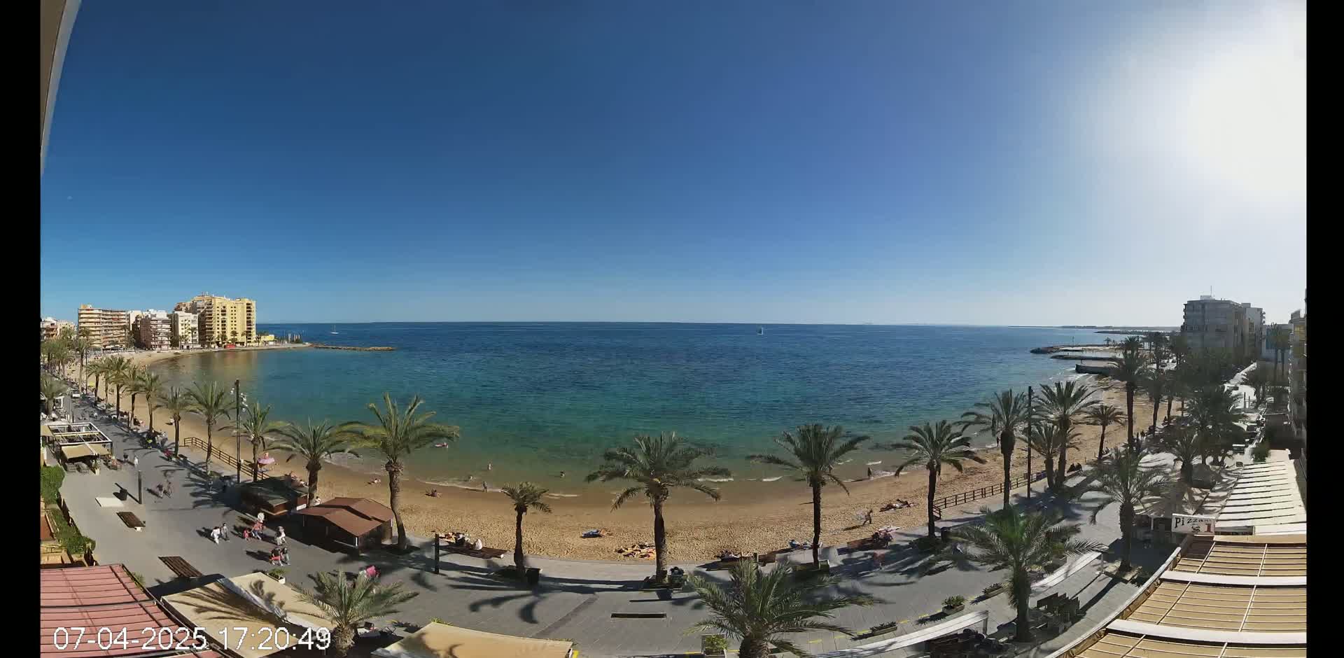 A sunny day at a sandy beach with palm trees lining a promenade, overlooking a calm, clear ocean with buildings visible in the distance.