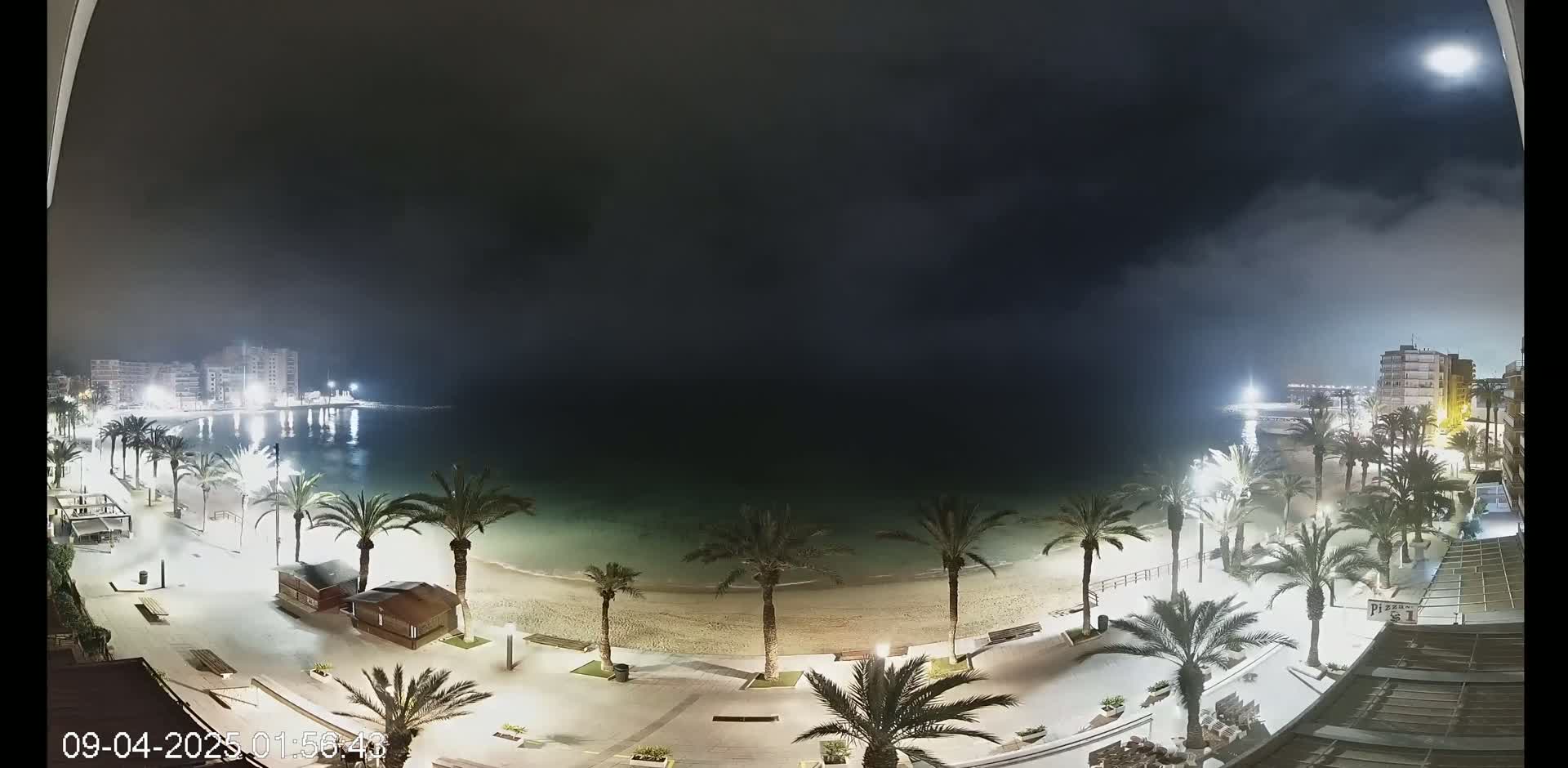 A nighttime view of a beach at the edge of a town under a mostly cloudy sky with a bright moon.