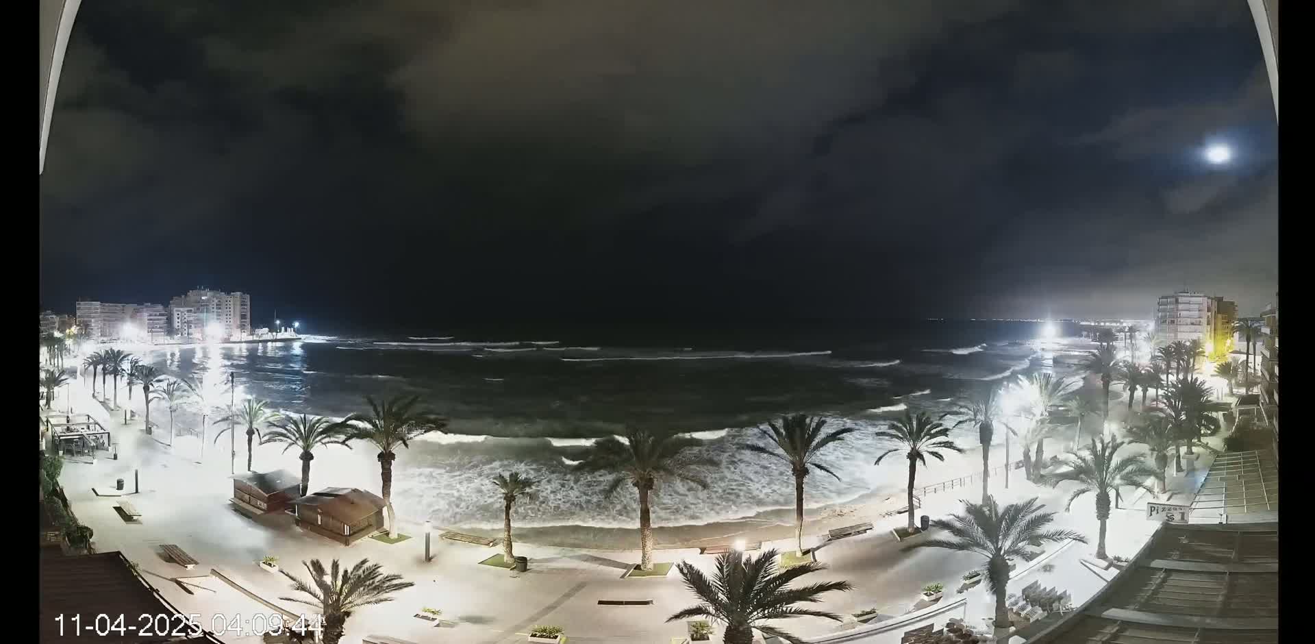 A nighttime view of a beach with moderately rough waves under a partly cloudy sky, palm trees lining a paved beachfront walkway, and buildings in the background.