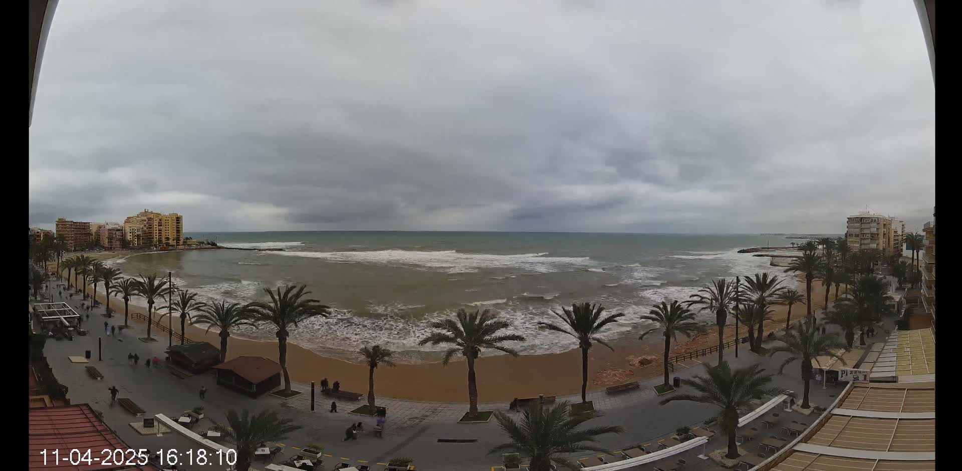 A mostly cloudy day shows a sandy beach with palm trees lining a paved walkway, buildings in the background, and a somewhat choppy ocean with waves breaking near the shore.
