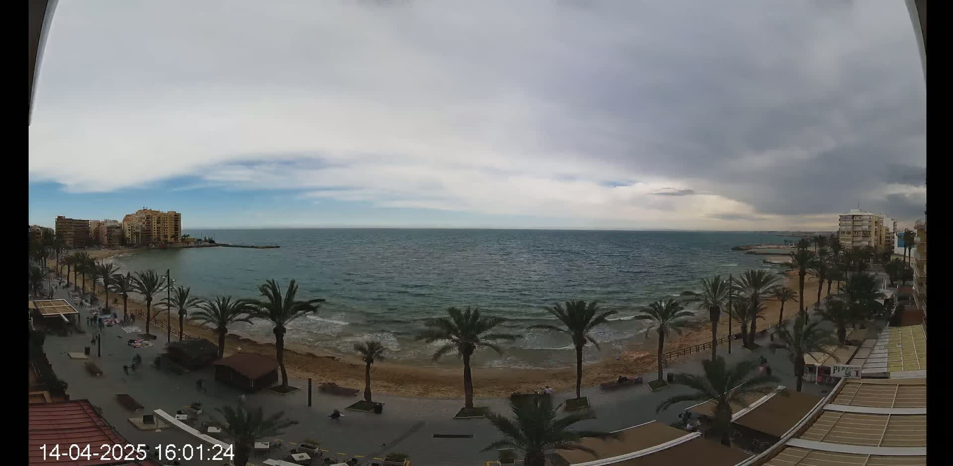 An overcast sky hangs over a sandy beach lined with palm trees and buildings, with a calm sea visible in the background.