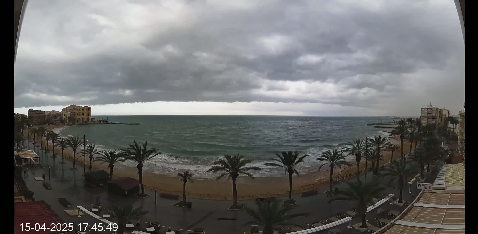 A beachside promenade with palm trees is partially flooded under a stormy, overcast sky, with waves crashing on the shore.