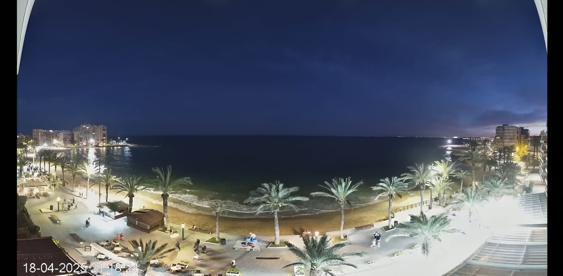 A nighttime view of a beach with palm trees, buildings, and a few people walking along the shore under a clear, dark blue sky.