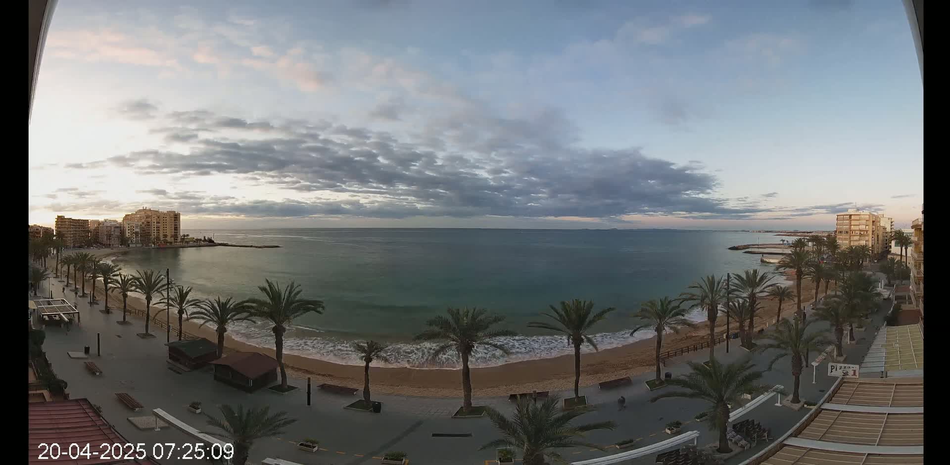 A sandy beach lined with palm trees and buildings sits on a calm sea under a cloudy sky.