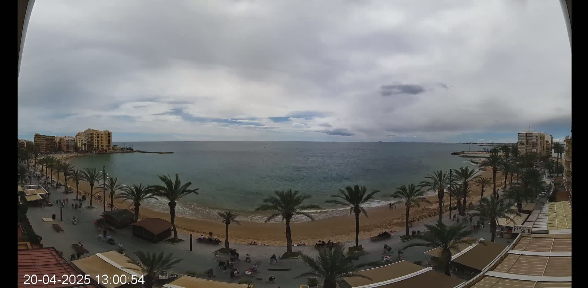 An overcast day reveals a sandy beach lined with palm trees, bordered by buildings and a promenade with people strolling along the shore next to calm, blue water.