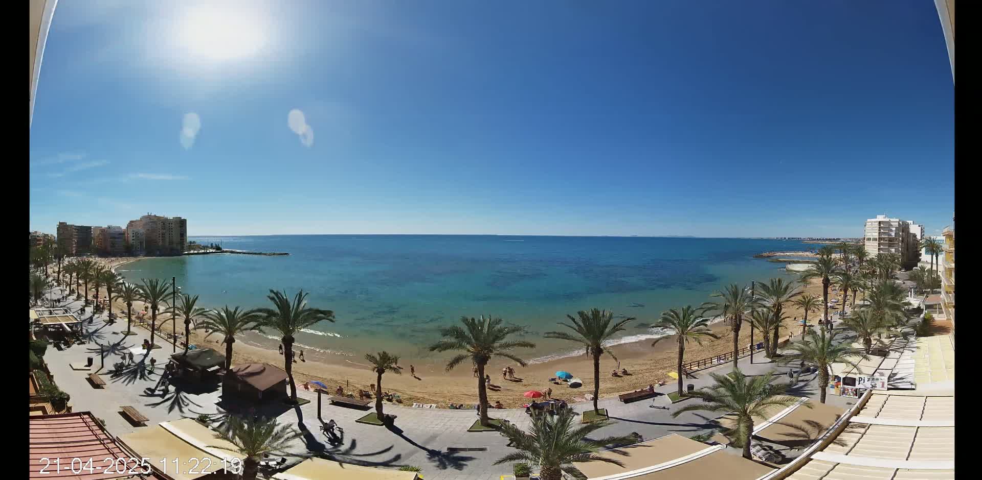 A sunny day at a sandy beach, lined with palm trees and buildings, overlooks a calm, turquoise sea.