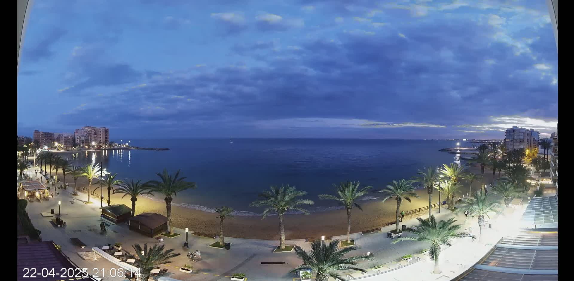 A twilight view shows a sandy beach lined with palm trees and buildings, overlooking a calm, dark-blue sea under a partly cloudy sky.