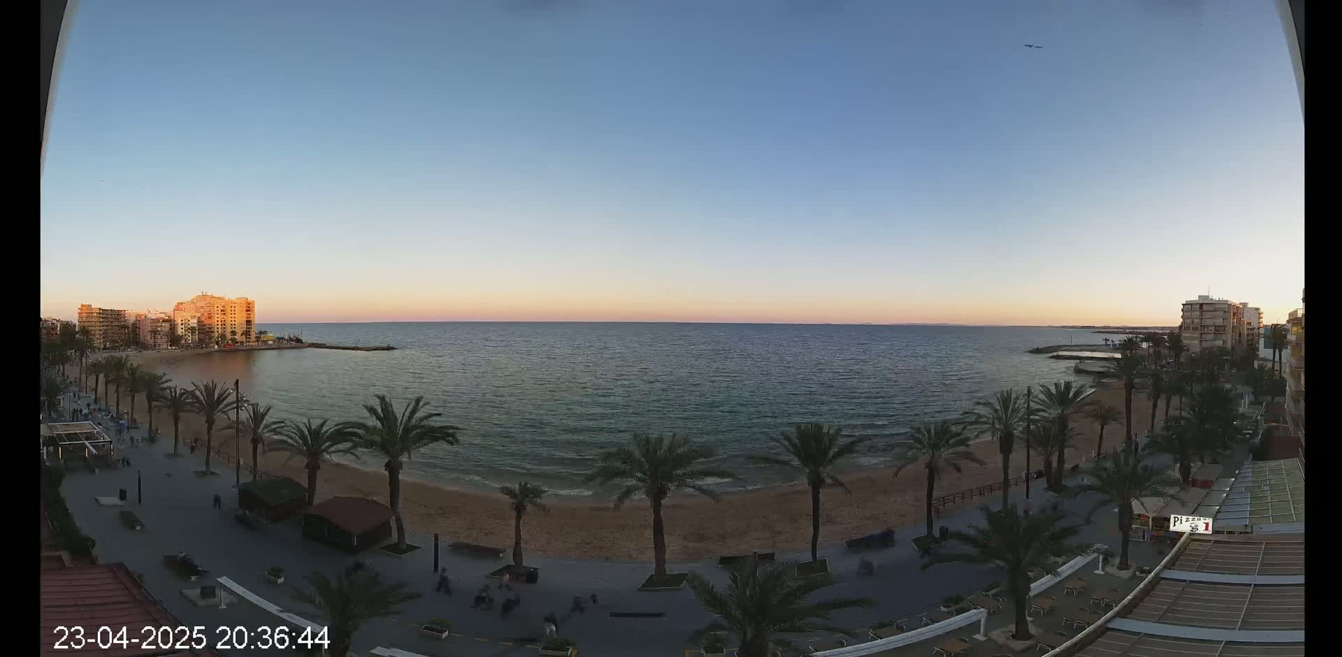 A calm sea at sunset is viewed from a high vantage point overlooking a sandy beach lined with palm trees and buildings.