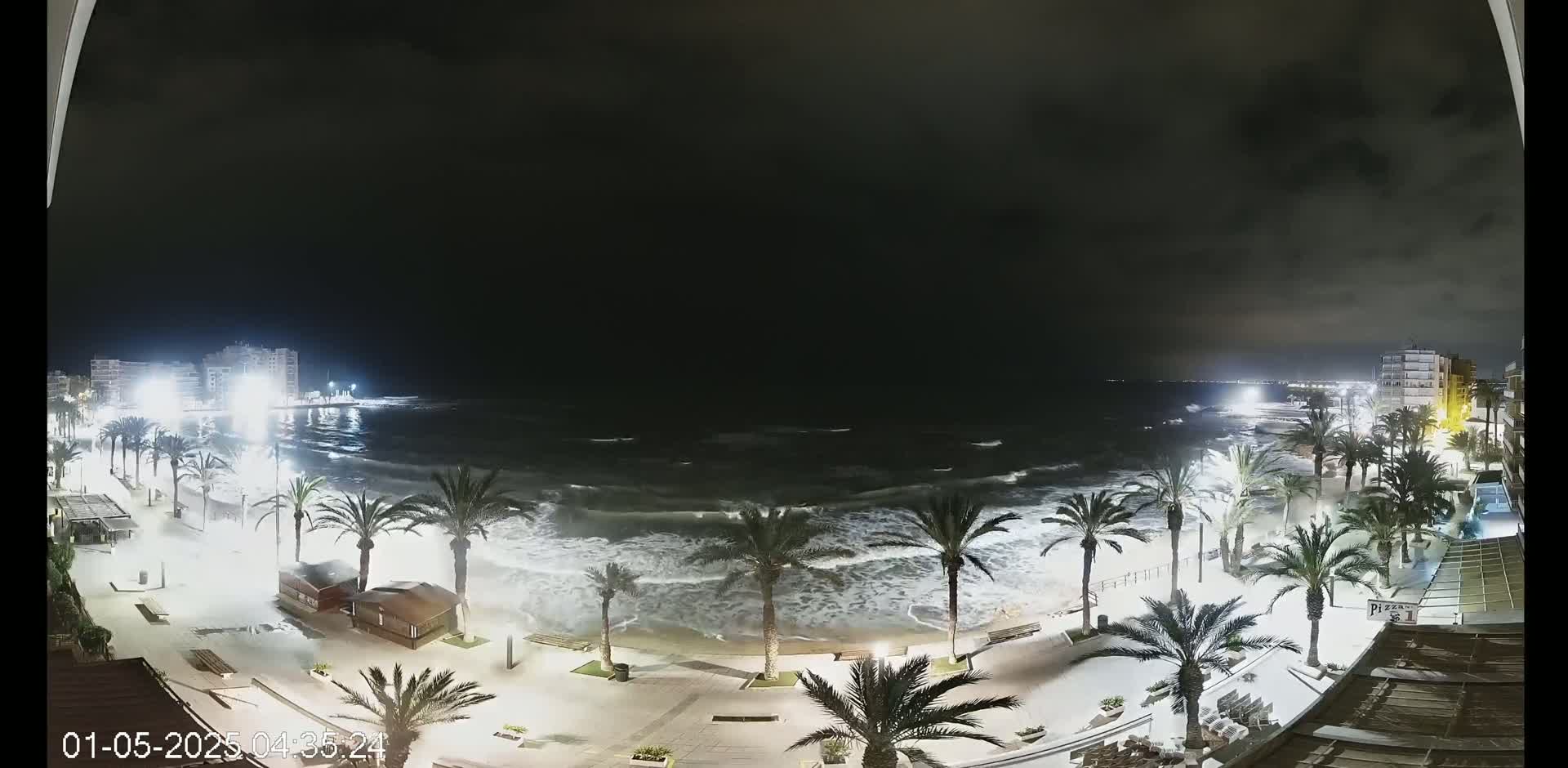 A nighttime view of a beach with palm trees and buildings along a promenade, with choppy waves crashing on the shore under a dark, cloudy sky.