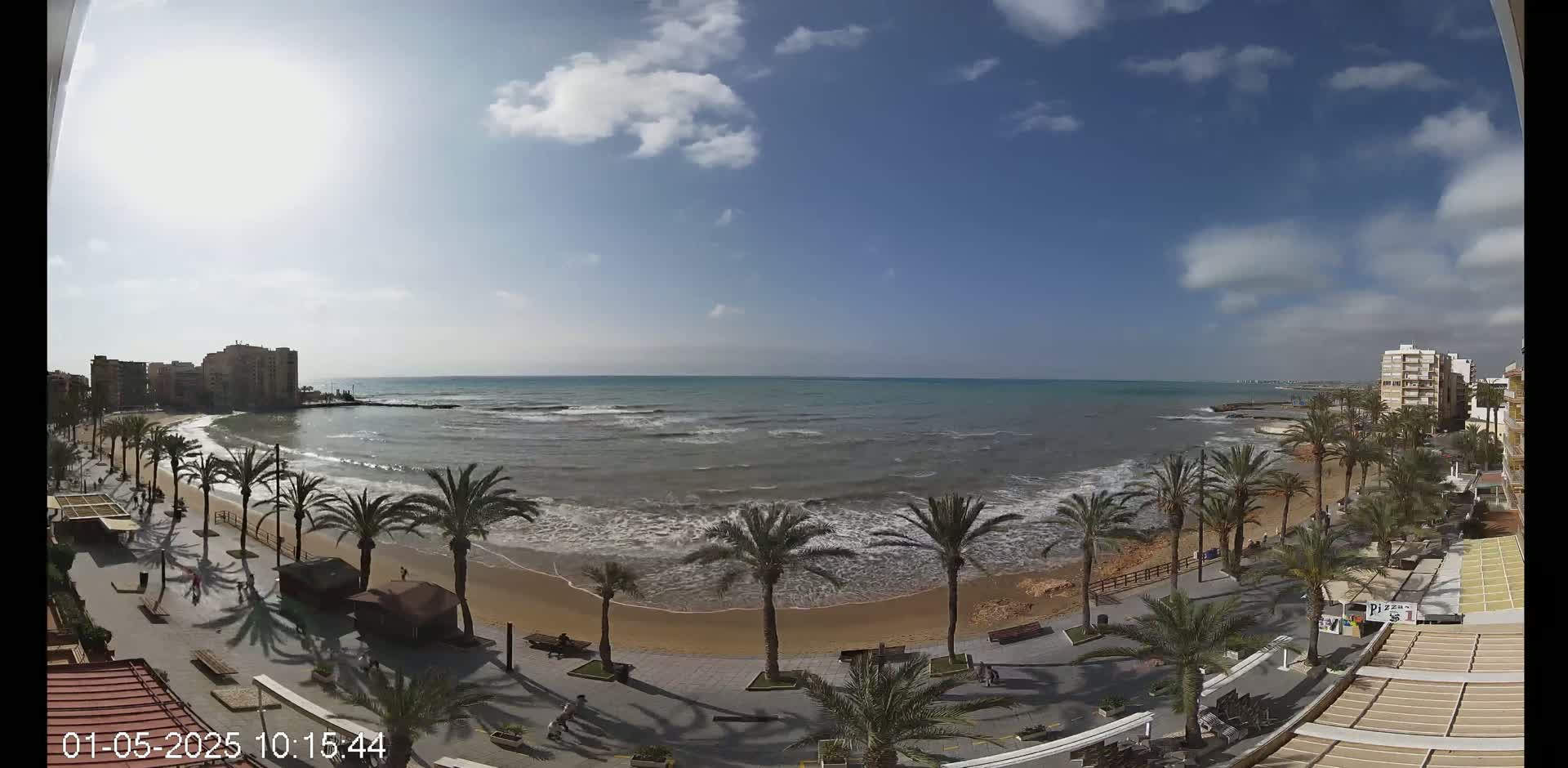 A mostly sunny day at a beach with palm trees lining a paved walkway next to a sandy shore and gently rolling waves.