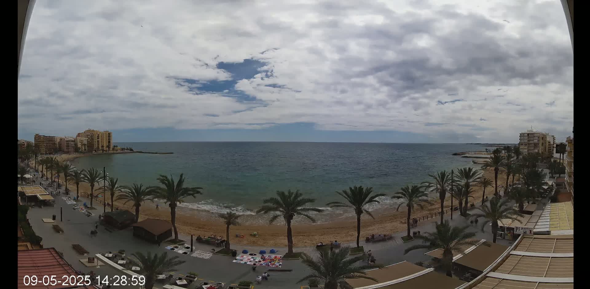 A mostly cloudy day overlooks a sandy beach with palm trees, a boardwalk, and buildings along a calm ocean.