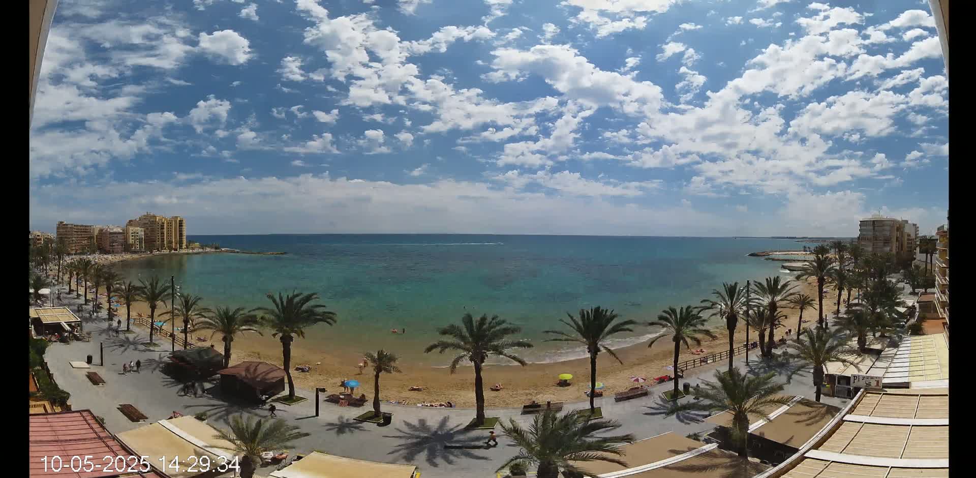 A partly sunny day shows a sandy beach lined with palm trees and buildings along a calm, turquoise ocean.