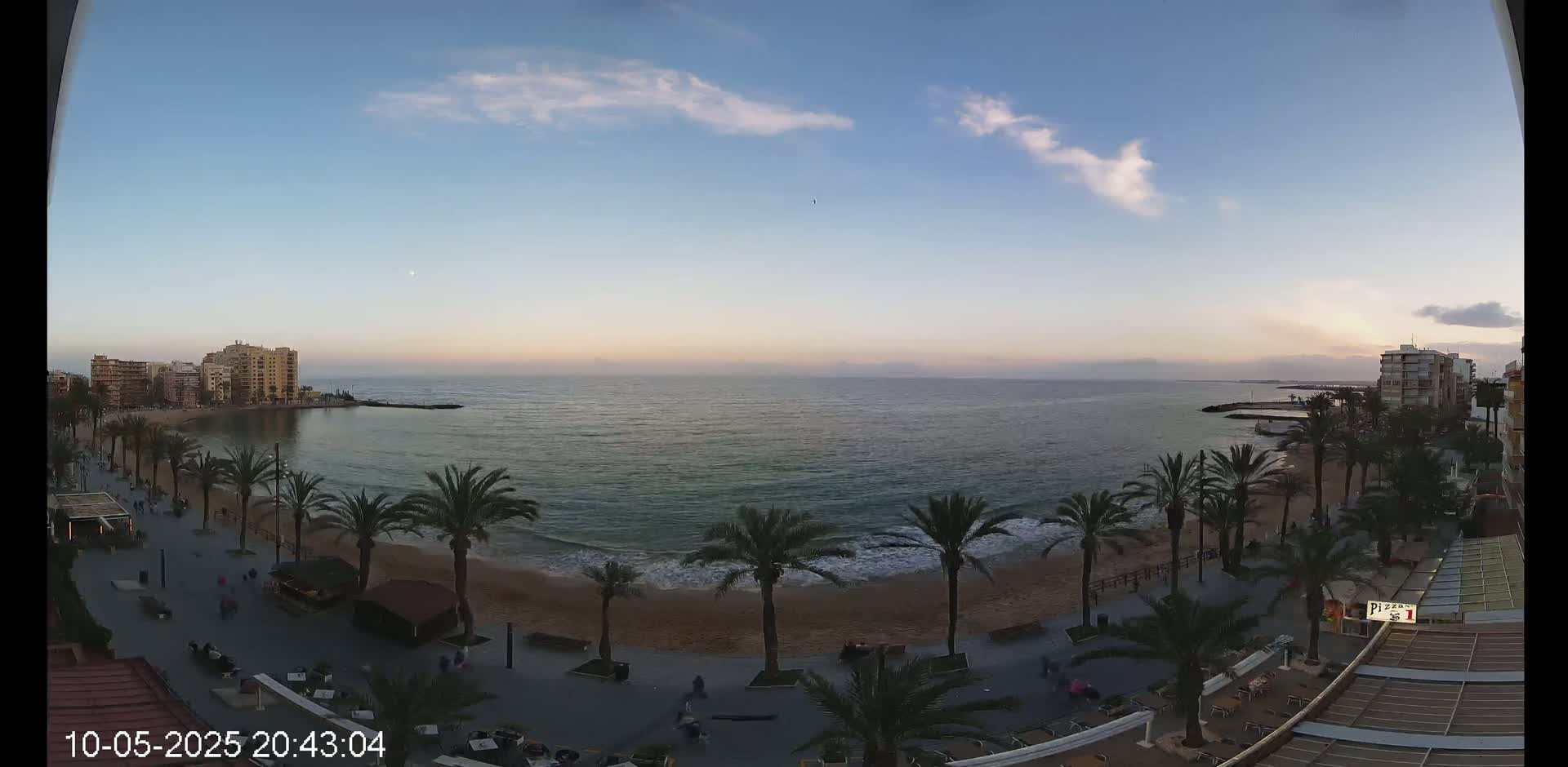 A mostly clear evening sky overlooks a sandy beach lined with palm trees and buildings, with calm ocean waves lapping the shore.